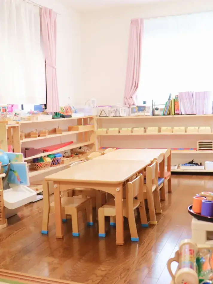 木製の低い机と椅子、モンテッソーリ教具の棚が並ぶ明るい保育室